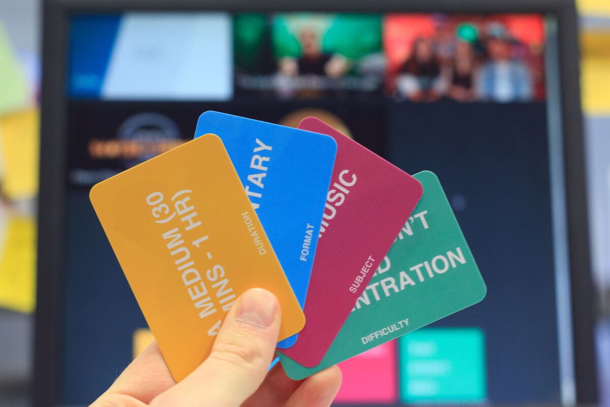 A hand holding three brightly coloured playing cards in front of a TV screen – the cards say things like “A documentary” “About music” “less than half an hour”