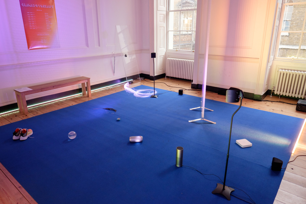 A gallery space with a blue rug, upon which are a series of objects, including an amazon alexa, a crystal globe, a pair of sports shoes, a bunch of fibre optics, a bluetooth speaker and a standing lamp