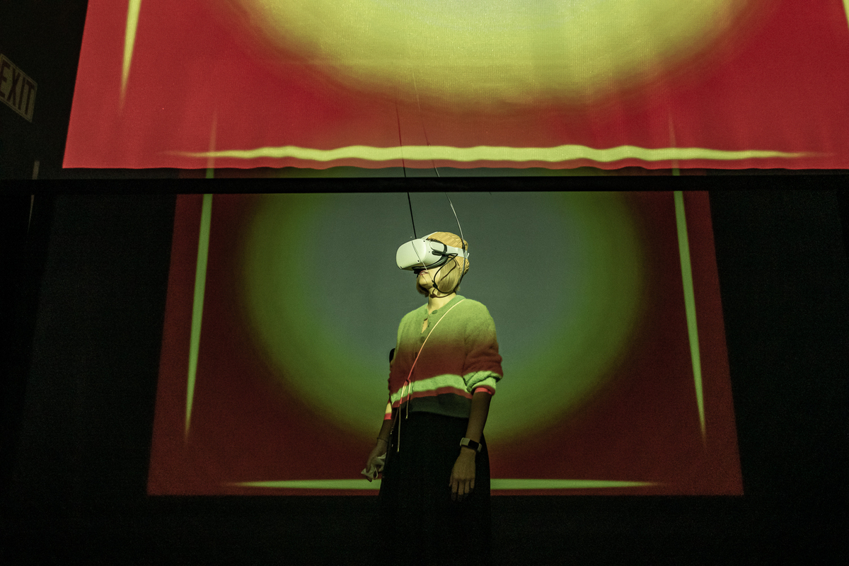 A woman wearing a VR headset, standing in a darkened space behind a translucent screen onto which are being projected green and red geometric shapes